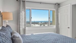 Un dormitorio con un ventanal grande, una cama con colcha estampada, una lámpara de noche y vistas a la playa.