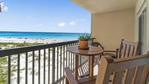 Un balcón con vista a la playa, una mesa redonda de madera con una planta en maceta y dos sillas de madera.