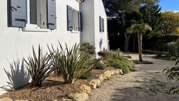 A white building with a tiled roof, a gravel pathway, and various plants.