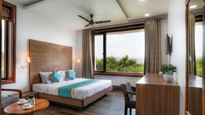 A modern hotel room with a large bed, a desk, a chair, a ceiling fan, and a large window offering a view of greenery. - Shivani Foods And Resort (Ranikhet)