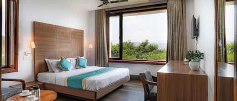Una habitación de hotel moderna con una cama grande, un escritorio, una silla, un ventilador de techo y un ventanal con vistas a la vegetación.