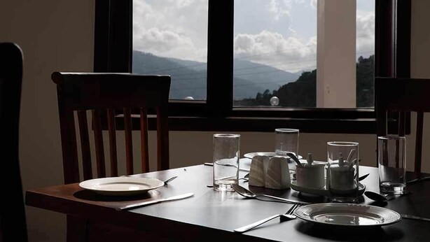 A dining table with chairs, a plate, cutlery, and glasses, set against a window with a mountain view. - Shivani Foods And Resort (Ranikhet)