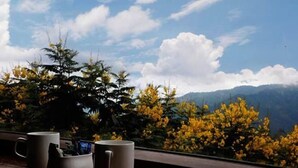 A window with three mugs on a wooden ledge, overlooking a mountain landscape with yellow flowering trees and a clear sky. - Shivani Foods And Resort (Ranikhet)