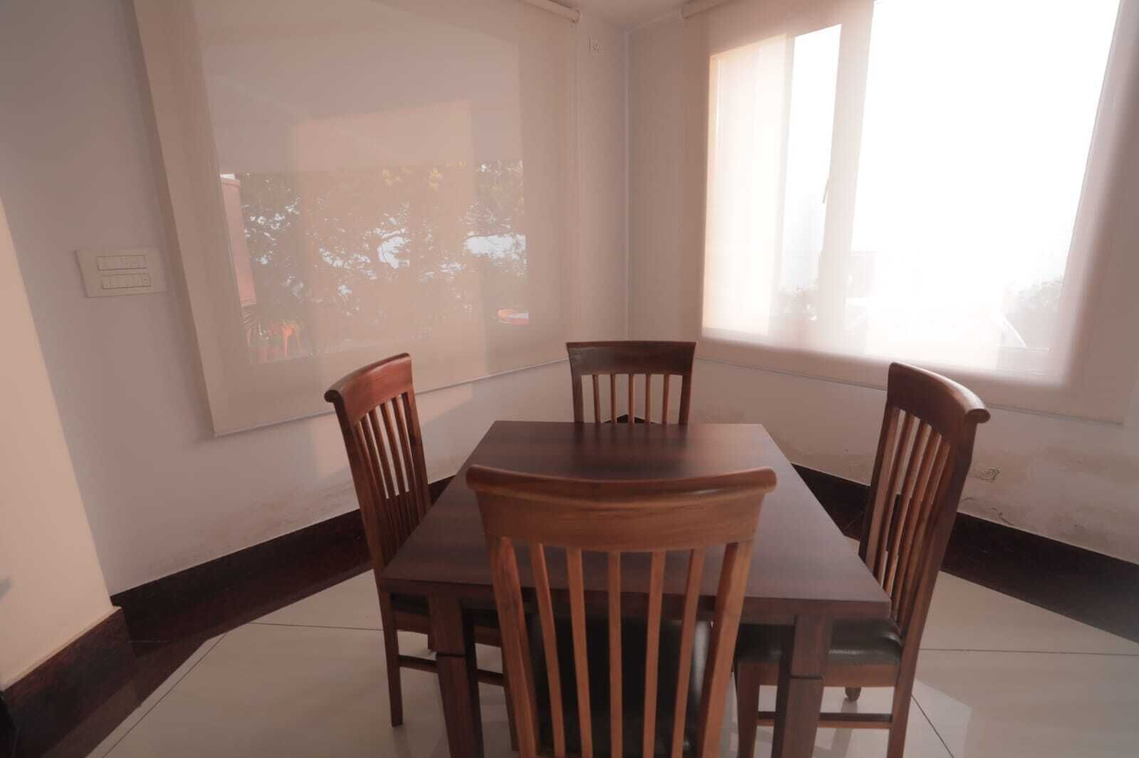 A dining area with a rectangular table and four wooden chairs, a large window with blinds, and a projection screen on the wall.