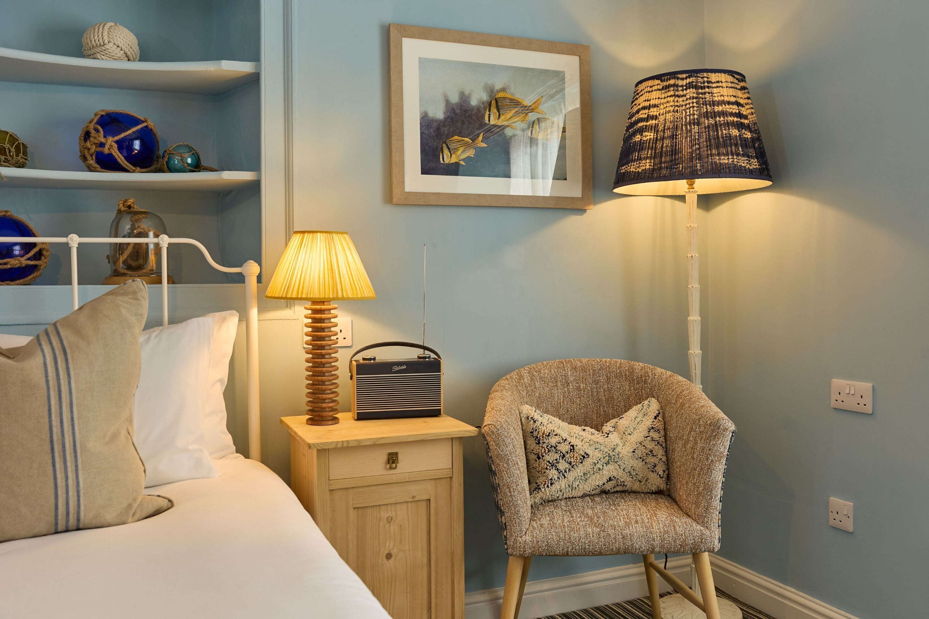 A neatly arranged bedroom with a bed, a chair, a bedside table with a lamp, a radio, and decorative items on shelves.