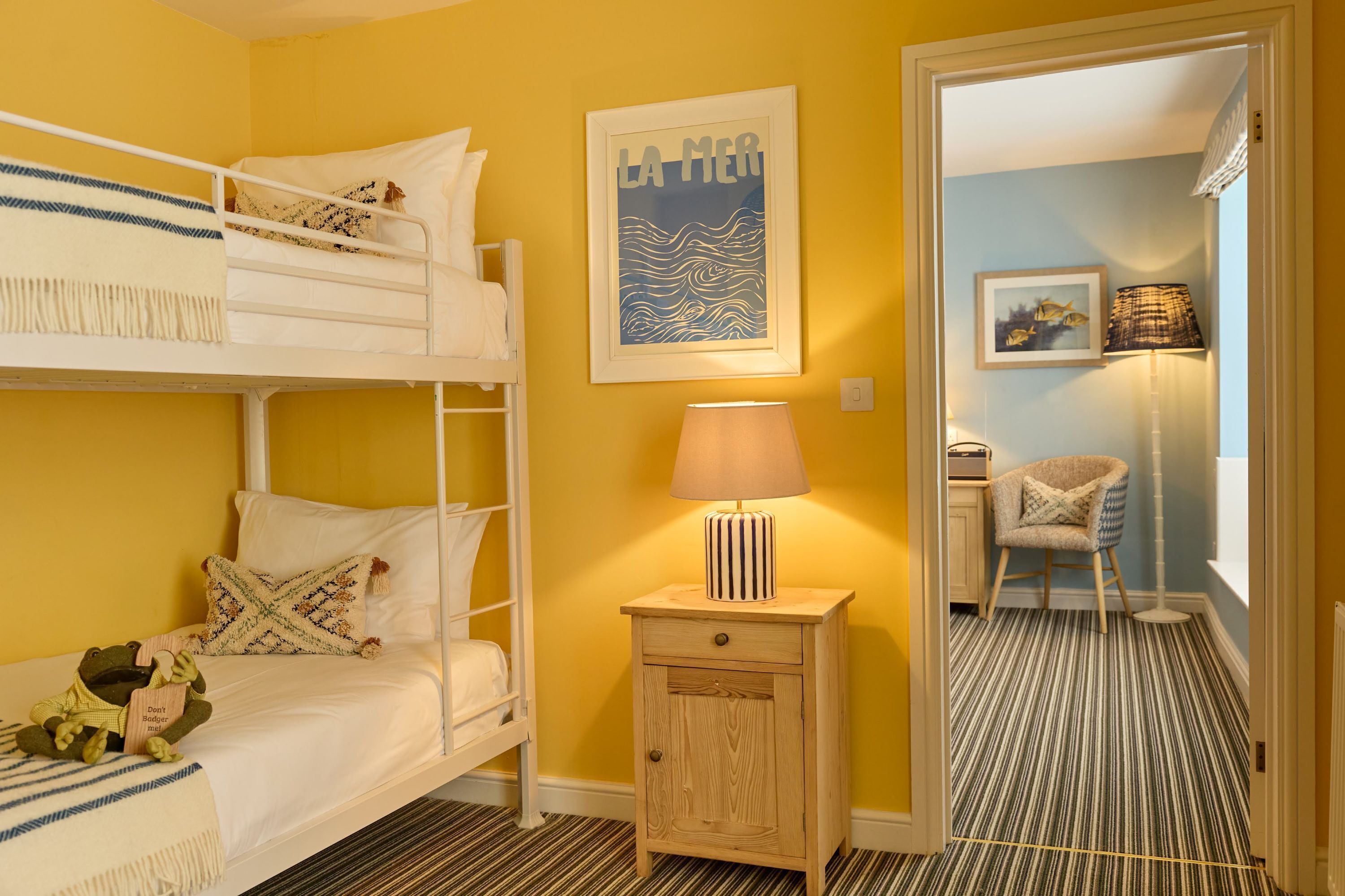Una habitación con una cama individual, una mesita de noche con una lámpara, un cuadro en la pared y una silla en la habitación contigua.