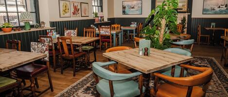 A cozy restaurant interior with wooden tables and chairs, potted plants, and artwork on the walls.