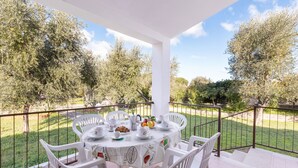 A covered outdoor dining area with a table set for four, surrounded by white chairs, and a view of a grassy garden with trees.