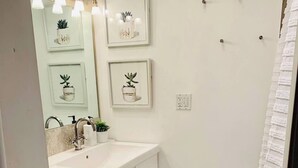 Un baño con lavamanos blanco, inodoro y zona de ducha. Hay cuadros enmarcados en la pared y un perchero.