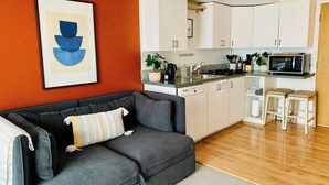Una sala de estar moderna con un sofá gris, un cuadro abstracto enmarcado y una cocina con alacenas blancas y un microondas.