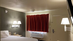 A hotel room with a bed, two bedside lamps, a window with red curtains, a door, and a wall-mounted air conditioner. - AMERICAN INN (Temple)