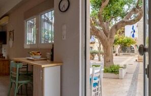 Una habitación con una mesa de madera, un cuenco de frutas y una silla. Hay una ventana con barrotes y vistas al exterior.