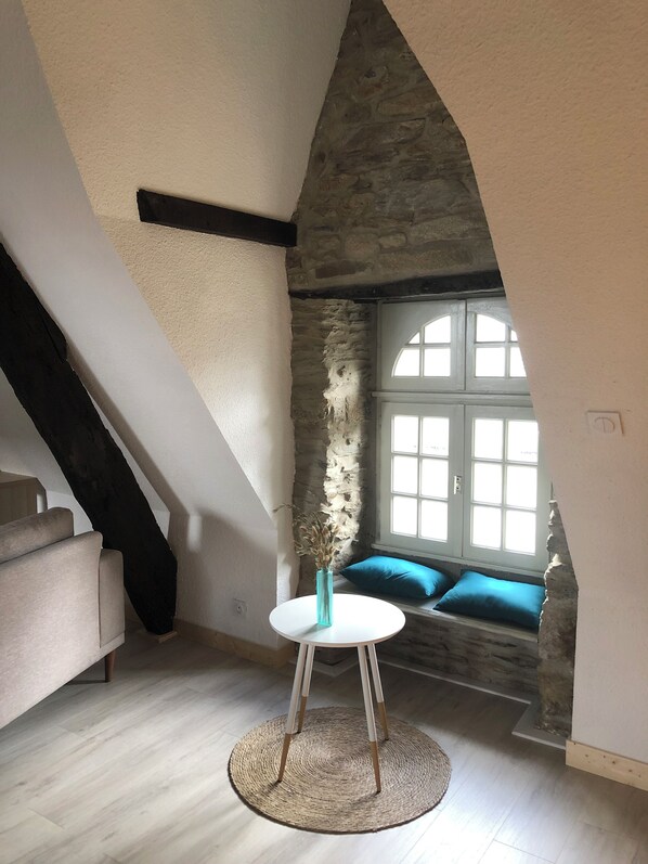 Une pièce mansardée chaleureuse avec un mur en pierre, une table ronde blanche ornée d’un vase et un banc bleu rembourré près d’une fenêtre.