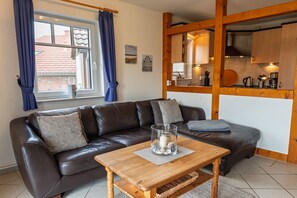 Una sala de estar con un sofá de cuero marrón, una mesa de centro de madera, y una zona de cocina visible a través de una partición de madera abierta.