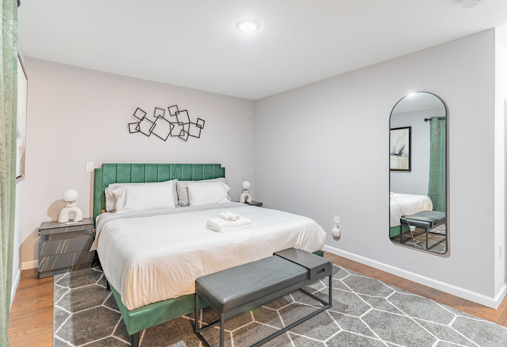 A bedroom with a bed, bedside tables, a bench, a mirror, and a geometric wall art piece.