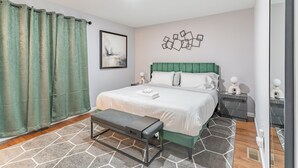 A modern bedroom with a green upholstered headboard, a large bed with white linens, a gray bench, and a geometric wall decoration. - Comfy Unit Loft 15 min to EWR (Newark)
