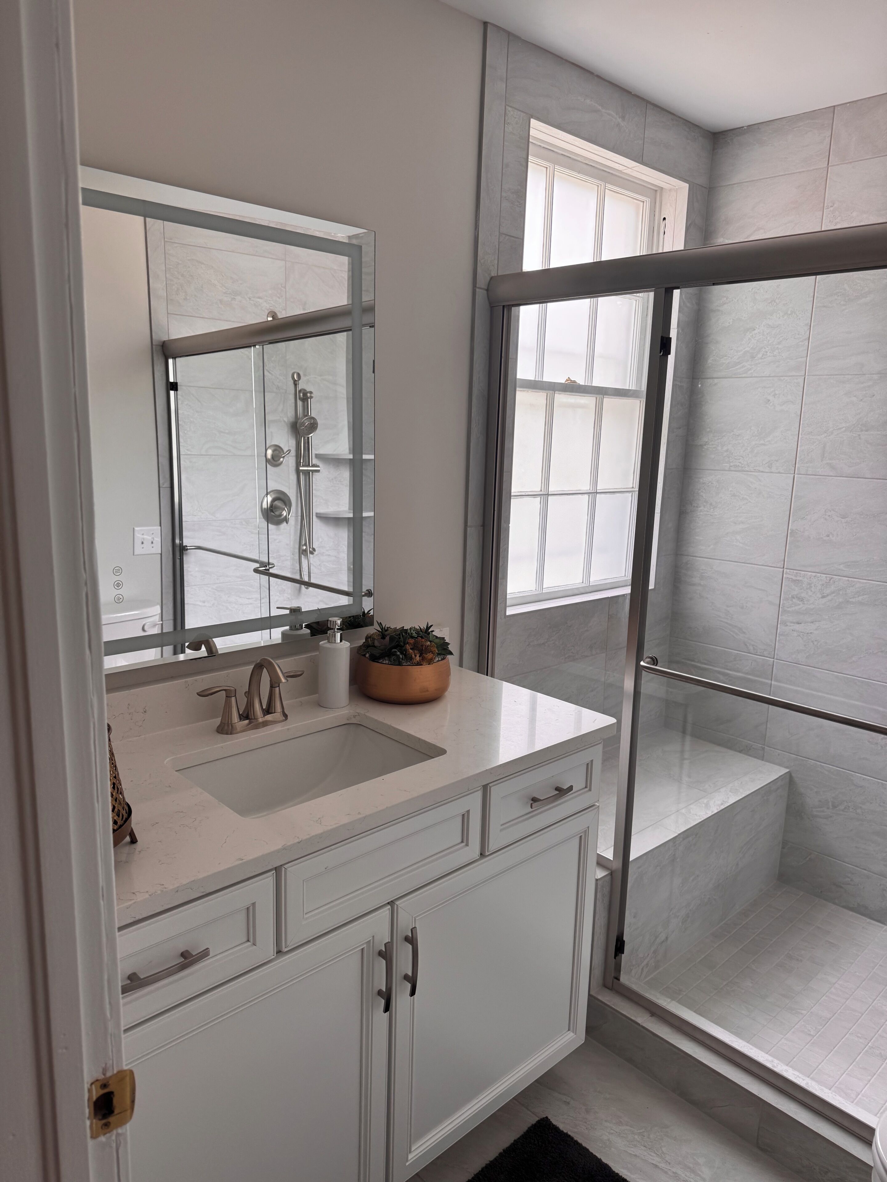 A bathroom with a white vanity, a large mirror, a shower with a glass door, and a window providing natural light.