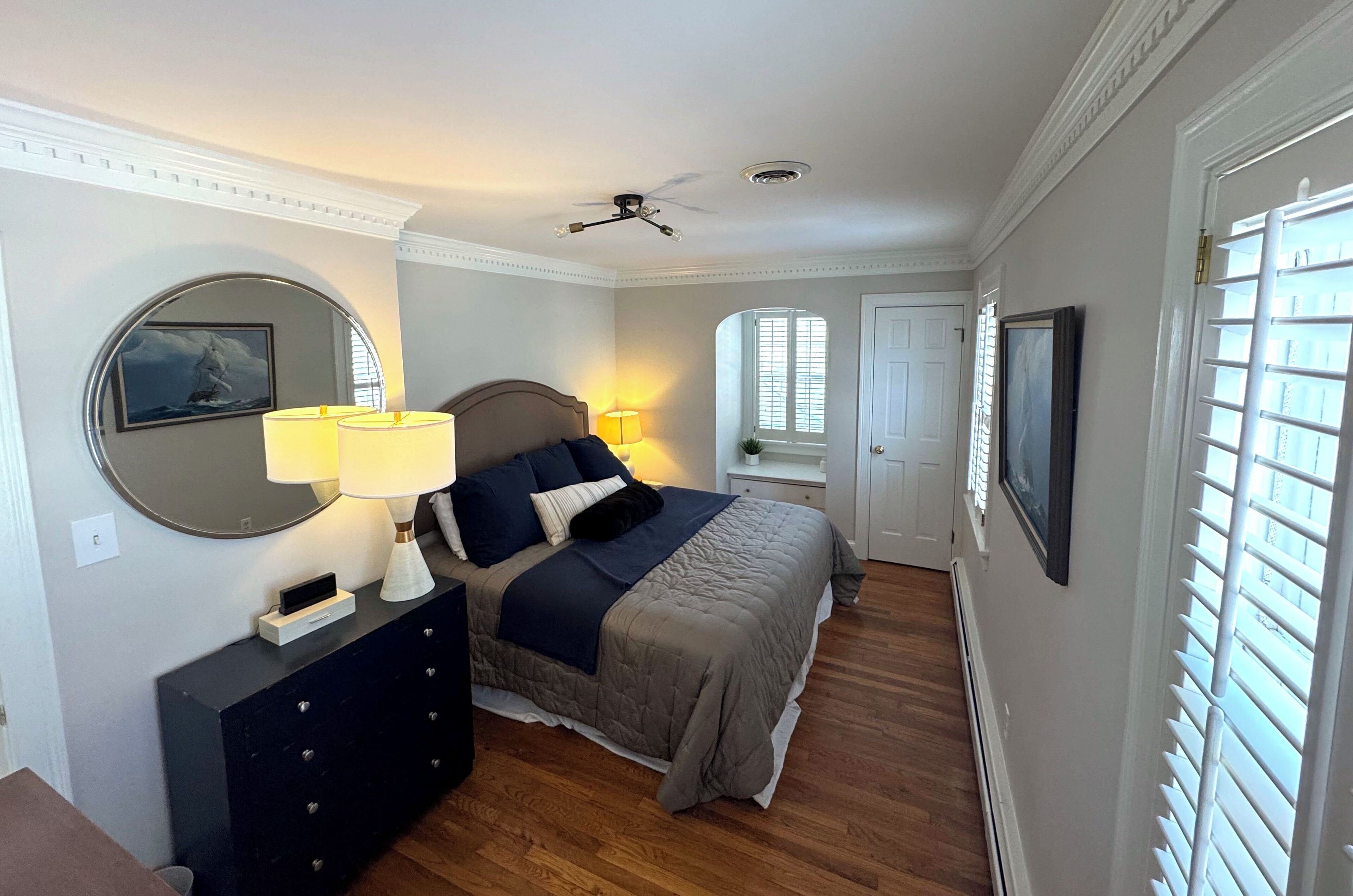 Une chambre à coucher comprenant un lit, des lampes de chevet, un ventilateur de plafond, une commode, un miroir et une fenêtre avec des stores.