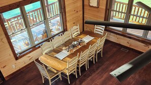 A wooden dining area with a table set for six, large windows with a view of a wooden deck, and a wooden floor.