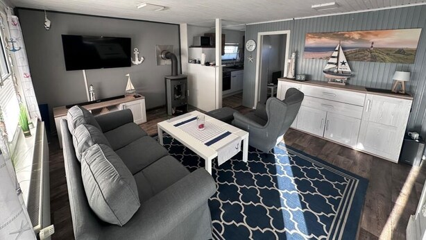 A modern living room with a sofa, coffee table, and a TV. There is a kitchen area with cabinets and a microwave. A painting of a sailboat is on the wall. - NEW! Vacation home Nordlicht (Südbrookmerland)
