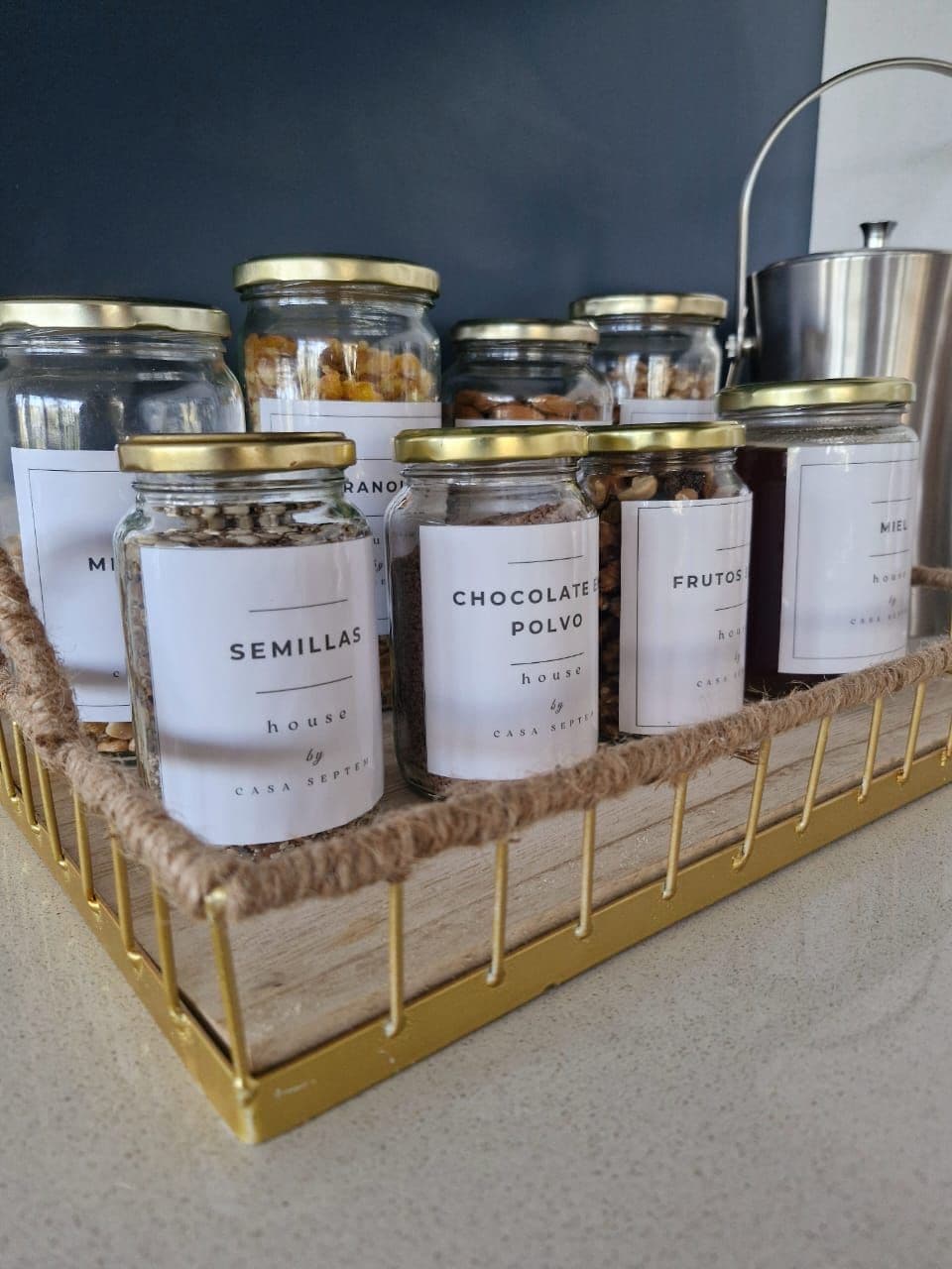 A display of glass jars with metal lids, each labeled with different contents like "Semillas" and "Chocolate Polvo," arranged on a golden wire rack.