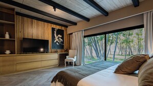 A modern bedroom with a large bed, a TV mounted on a wooden wall, a chair, and a view of trees through large windows. - House by Casa Septem (Tunuyan)