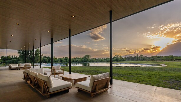 A spacious outdoor area with multiple seating options, large windows offering a view of a lake and sunset, and a modern architectural design. - House by Casa Septem (Tunuyan)