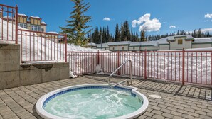 A hot tub with a metal railing, surrounded by a brick patio, with a snowy landscape and buildings in the background.