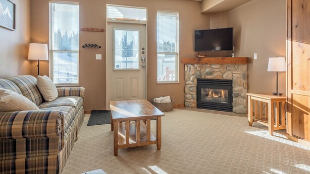 A cozy living room with a plaid sofa, a wooden coffee table, a stone fireplace, and a television mounted above it.