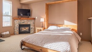 A bedroom with a stone fireplace, a television mounted above it, a bed with a wooden headboard, and a window with blinds.
