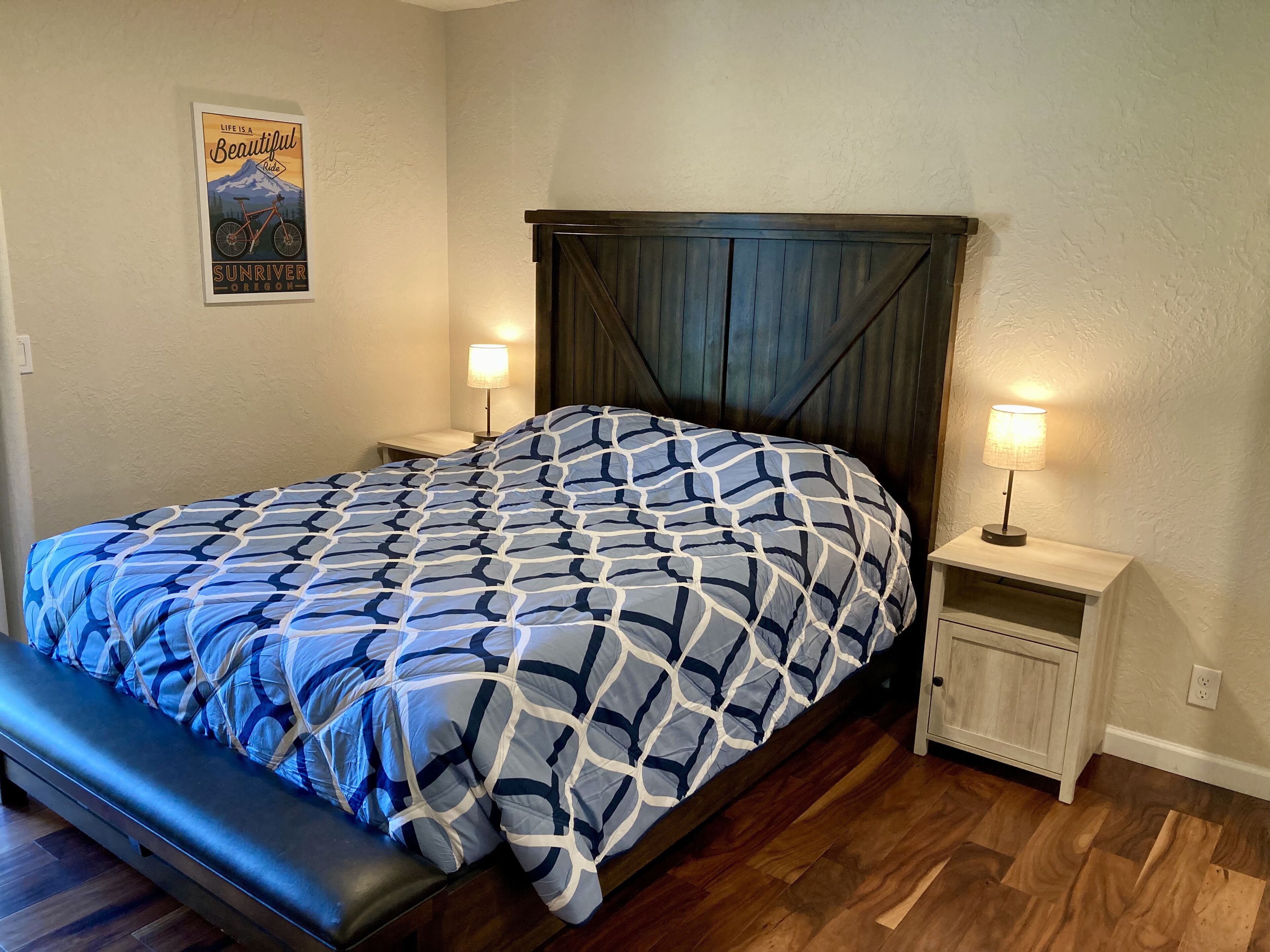 A bedroom with a bed, two bedside tables, a bench, a poster on the wall, and a wooden floor.