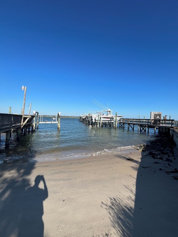 Un muelle se extiende sobre aguas tranquilas con barcos amarrados, una playa de arena y un cielo azul despejado.