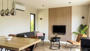 A modern living room with a wooden dining table, a black sofa, and a TV mounted on a wooden wall.