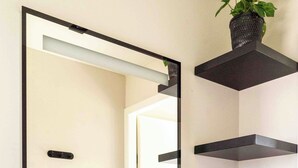 A modern bathroom with a white sink, black faucet, and a mirror above it. There are black floating shelves and a potted plant on the wall.