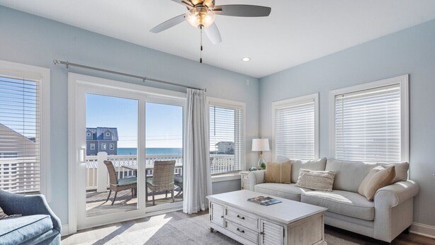 A spacious living room with a large sliding glass door leading to a balcony, a white coffee table, a sofa, and a ceiling fan. - Topsail Treasure: Elevator| Pool| Steps from Beach (Surf City)