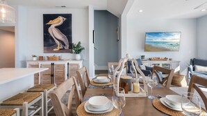 A dining area with a long table set for six, surrounded by wooden chairs. There is a painting of a pelican on the wall, a plant, and a framed picture of a beach scene. - Topsail Treasure: Elevator| Pool| Steps from Beach (Surf City)