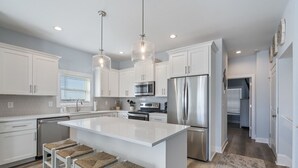 A modern kitchen with white cabinetry, stainless steel appliances, and a large island with bar stools. - Topsail Treasure: Elevator| Pool| Steps from Beach (Surf City)