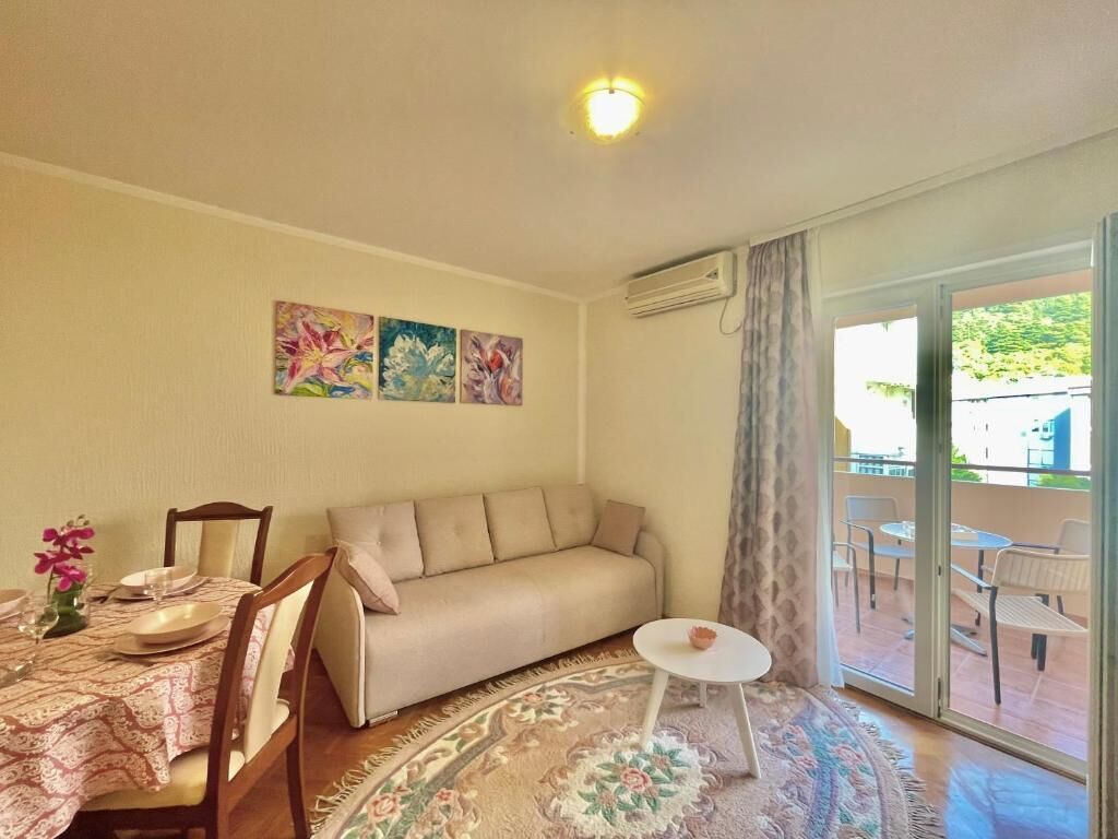 Una sala de estar con un sofá, una mesa de comedor y sillas. Hay tres cuadros en la pared, una mesita redonda y vistas al exterior a través de un ventanal.