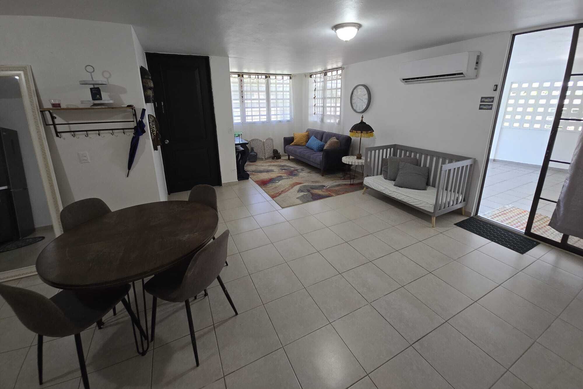 Una sala de estar con una mesa de comedor, sofá y una cuna para bebé. Hay una puerta que conduce a otra habitación y un equipo de aire acondicionado en la pared.