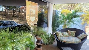 Un espace extérieur abrité, aménagé avec des meubles en osier, des plantes en pot et un panneau affichant des armoiries.