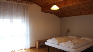Un dormitorio con techo de madera, una cama con ropa de cama blanca, una banca y una ventana con cortinas traslúcidas.