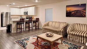 Una cocina moderna con gabinetes blancos, un refrigerador de acero inoxidable y una barra con taburetes. Hay una mesa de comedor con un cuenco decorativo y una vela. La sala de estar tiene sofás a rayas y una alfombra colorida.