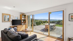 Ein Wohnzimmer mit Fernseher, Bücherregal und einer großen Schiebetür, die auf eine Terrasse mit Blick ins Grüne führt.