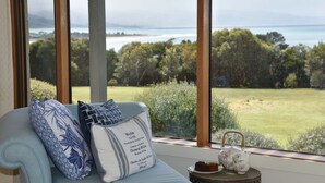 Ein Zimmer mit Blick auf ein grünes Feld und das Meer. Es gibt ein Sofa mit Zierkissen und einen kleinen Tisch mit Teekanne und einem Teller Essen.
