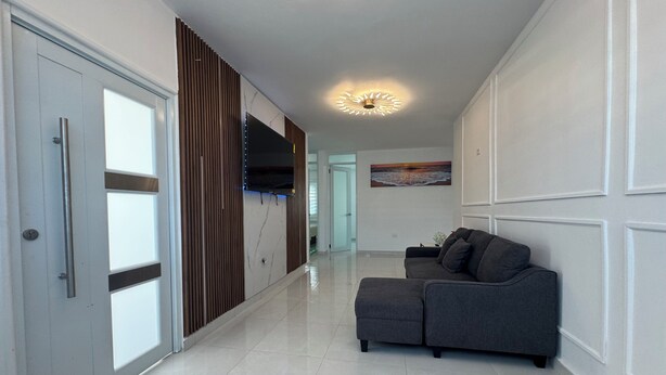 A modern hallway with a grey sectional sofa, a flat-screen TV mounted on a wooden panel, and a ceiling light fixture.