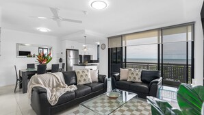 Ein modernes Wohnzimmer mit einem Glastisch, zwei schwarzen Ledersofas mit Palmenkissen, einem Essbereich mit weißem Tisch und Stühlen, einer Küche mit Edelstahlgeräten und einem großen Fenster mit Blick auf den Strand.
