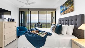 A hotel room with a bed, a blue armchair, a wooden dresser, and a balcony with a view of the ocean. - Belle Escapes 73 Rydges Plaza Esplanade with Ocean & City Views (Cairns North)