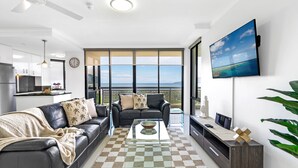 Ein modernes Wohnzimmer mit Schachbrettmuster-Boden, einer Couch mit Palmenmuster-Kissen, einem Couchtisch mit einer Schale, einem Fernseher an der Wand und einem Balkon mit Blick.