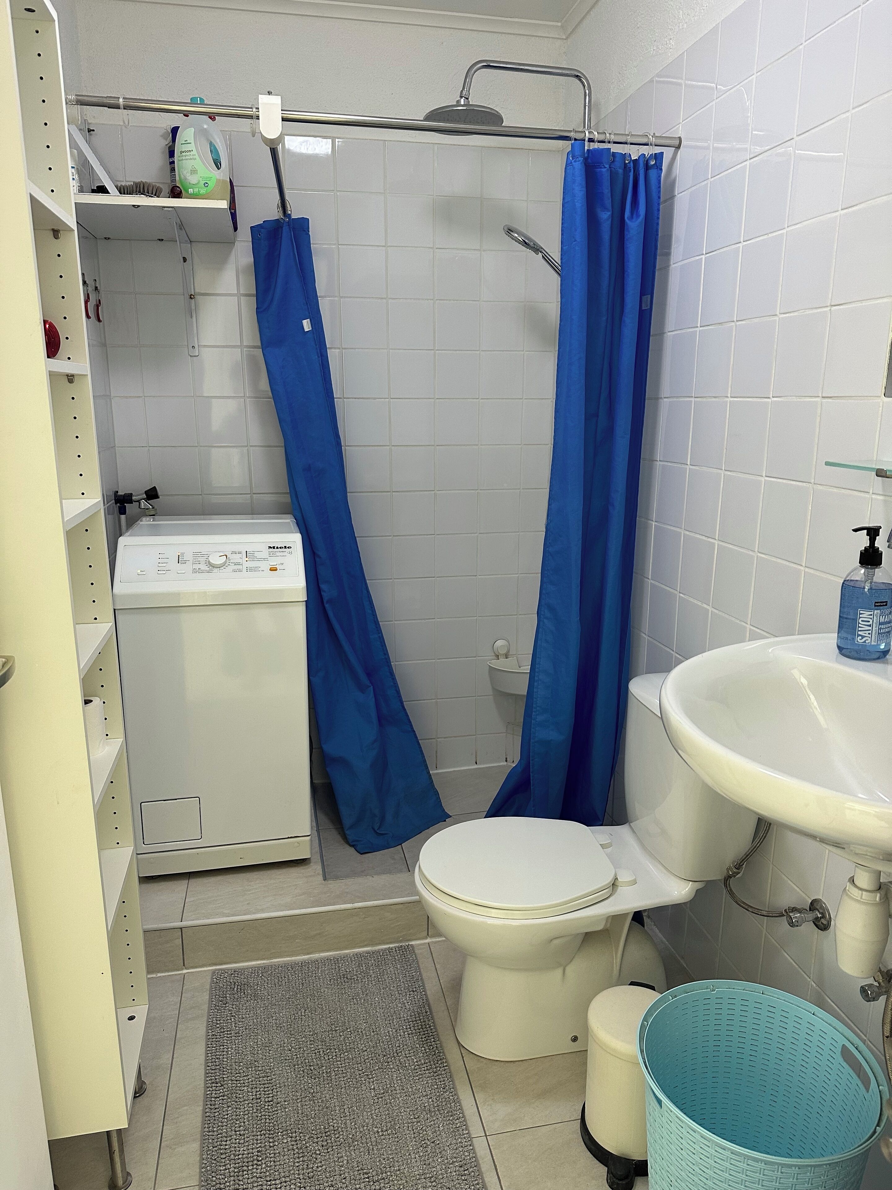 Un baño con inodoro, lavamanos, ducha con cortinas azules, lavadora y una cesta.