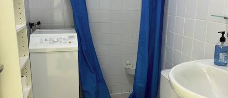 Un baño con inodoro, lavamanos, ducha con cortinas azules, lavadora y una cesta.